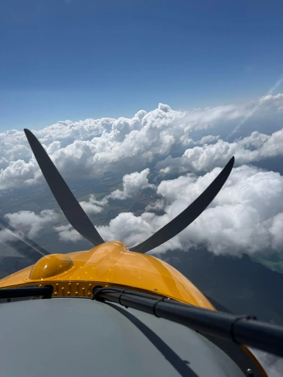 Blick aus dem Cockpit über die gelbe Flugzeugnase und den Propeller in eine hohe Wolkenlandschaft